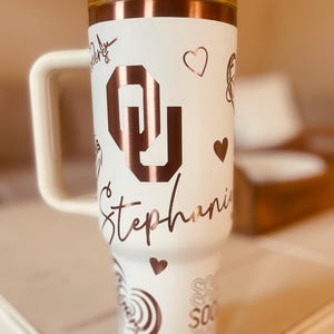 May include: White tumbler with a rose gold lid and handle. The tumbler has a rose gold design with the letter "O" and the name "Stephanie" on it.