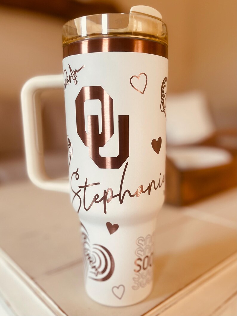 May include: White tumbler with a rose gold lid and handle. The tumbler has a rose gold design with the letter "O" and the name "Stephanie" on it.