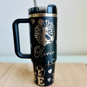 May include: Black stainless steel travel mug with a black handle. The mug has a gold glitter design with a cheerleader silhouette, pom poms, and the name "Olivia".