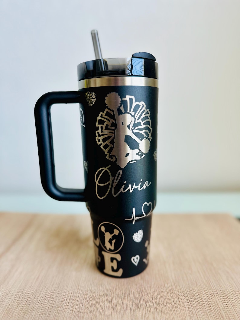 May include: Black stainless steel travel mug with a black handle. The mug has a gold glitter design with a cheerleader silhouette, pom poms, and the name "Olivia".