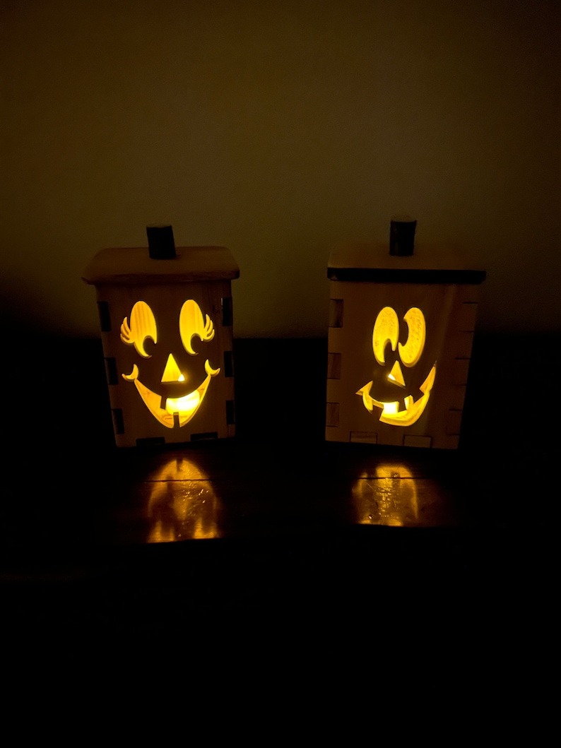 Puede incluir: Dos portavelas de madera con forma de calabaza con caras talladas y velas encendidas en su interior.