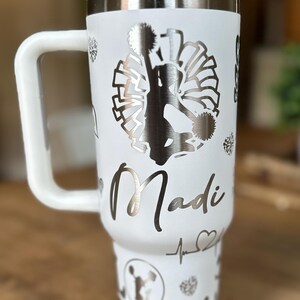 May include: White stainless steel travel mug with a black and white silhouette of a cheerleader holding pom-poms. The mug has a handle and the name "Madi" is engraved on the side.