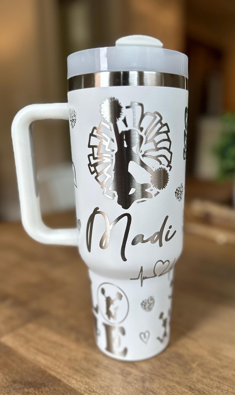 May include: White stainless steel travel mug with a black and white silhouette of a cheerleader holding pom-poms. The mug has a handle and the name "Madi" is engraved on the side.