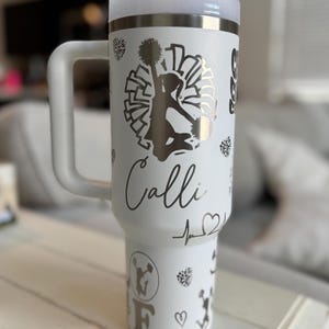 May include: White stainless steel tumbler with a black and white cheerleader design. The tumbler has a handle and a lid. The name "Calli" is written on the tumbler.