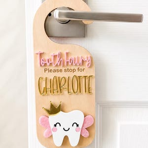 May include: A light-colored wooden door hanger featuring the pink 3D text Tooth Fairy, the brown text Please stop for, and the gold 3D name CHARLOTTE. Below, a white tooth icon with a gold crown and pink felt wings is attached to the wood.