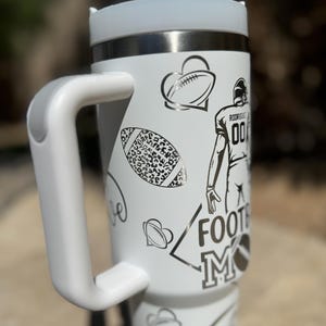 May include: White stainless steel travel mug with a black and white design featuring footballs, hearts, and the words "Football Mom".