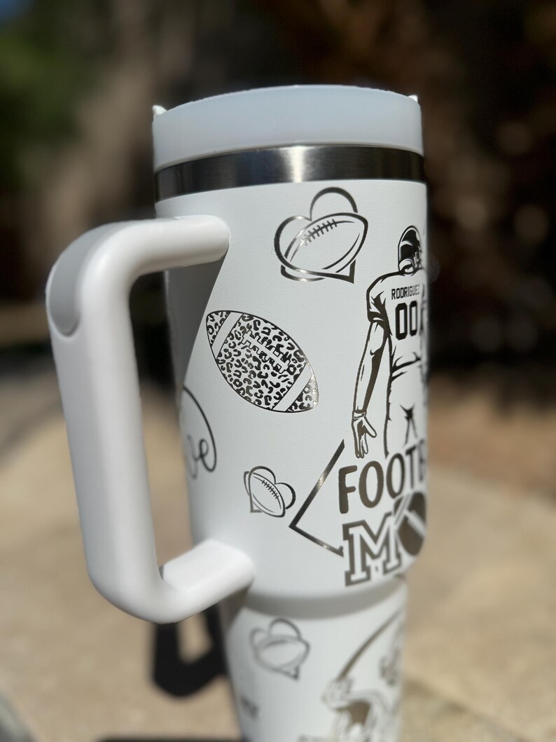 May include: White stainless steel travel mug with a black and white design featuring footballs, hearts, and the words "Football Mom".