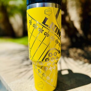 May include: Yellow stainless steel travel tumbler with a black lid. The tumbler has a football field design with a heart and the word "LIONS" on it.
