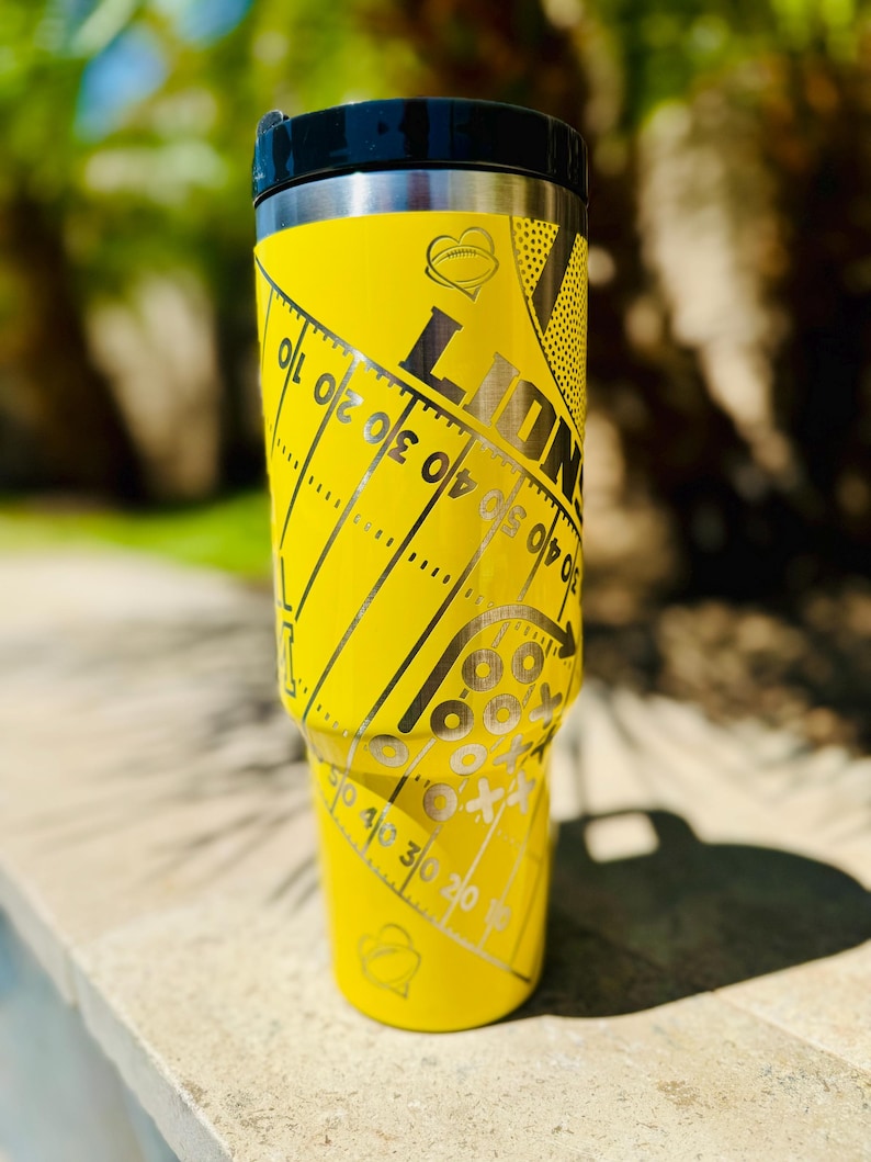 May include: Yellow stainless steel travel tumbler with a black lid. The tumbler has a football field design with a heart and the word "LIONS" on it.