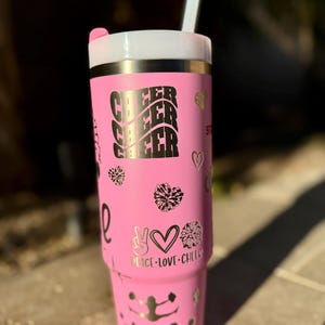 May include: Pink stainless steel tumbler with a white lid and a straw. The tumbler has a gold glitter design with the words "Cheer Cheer Cheer" and "Peace Love Cheer" on it. There are also several cheerleading pom-poms and a cheerleader silhouette on the tumbler.