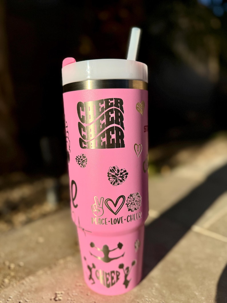 May include: Pink stainless steel tumbler with a white lid and a straw. The tumbler has a gold glitter design with the words "Cheer Cheer Cheer" and "Peace Love Cheer" on it. There are also several cheerleading pom-poms and a cheerleader silhouette on the tumbler.