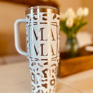 May include: White stainless steel travel mug with a handle. The mug has a leopard print design and the word "MAMA" in a gold frame.