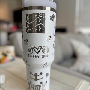 May include: White stainless steel tumbler with a silver design. The tumbler has a straw and features the words "Cheer Cheer Cheer", "Peace Love Cheer", and a cheerleader silhouette.