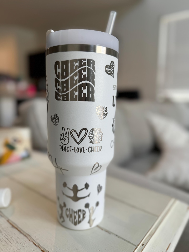 May include: White stainless steel tumbler with a silver design. The tumbler has a straw and features the words "Cheer Cheer Cheer", "Peace Love Cheer", and a cheerleader silhouette.