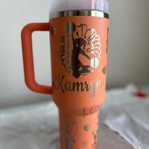 May include: A coral orange stainless steel tumbler with a lid and handle. The tumbler has a silver design of a cheerleader with pom-poms and the name "Kamryn" on the side.