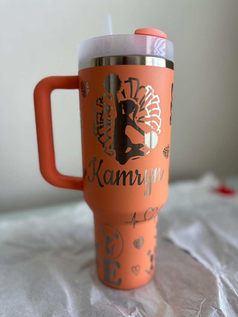 May include: A coral orange stainless steel tumbler with a lid and handle. The tumbler has a silver design of a cheerleader with pom-poms and the name "Kamryn" on the side.