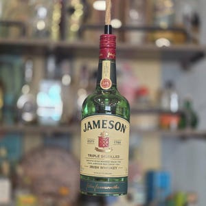 May include: A green glass Jameson Irish Whiskey bottle with a cream-colored label featuring the brand name, a red crest, and the text Triple Distilled. The bottle has a red screw cap with a thick tan rope extending upward from the center of the top.