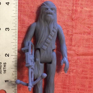 May include: A gray plastic action figure of Chewbacca from Star Wars. He is holding a blaster rifle and has a bandolier across his chest.