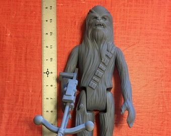 May include: A gray plastic action figure of Chewbacca from Star Wars, holding a blaster rifle. The figure is approximately 12 inches tall.