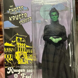 May include: Collectible action figure of a green-skinned woman in a black dress, packaged in a clear plastic case. The packaging features the text "The Ghosts of Kruger Street" and "Kruger Fest 2021".