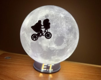 May include: A spherical lamp resembling the moon, with a black silhouette of a figure on a bicycle in front. The base of the lamp has the letters "E.T." in a glowing font. The lamp is on a wooden surface.