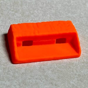 May include: A bright neon orange rectangular plastic component featuring a textured top surface and two small, centered rectangular cutouts along the front face. The object has a sloped front edge and a solid, block-like structure with defined corners.