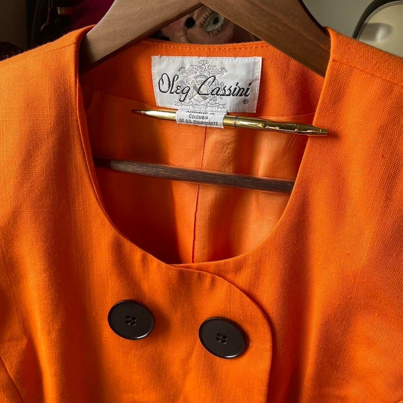 May include: Orange linen blazer with a scoop neck and two black buttons. The label reads "Oleg Cassini" with the words "Registered in Colombia" and "Design U.S. Patents" underneath.