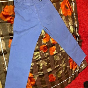 May include: A pair of blue jeans with a straight leg and frayed hem. The jeans are laid on a brown and orange floral patterned scarf, which is on top of a red rug.