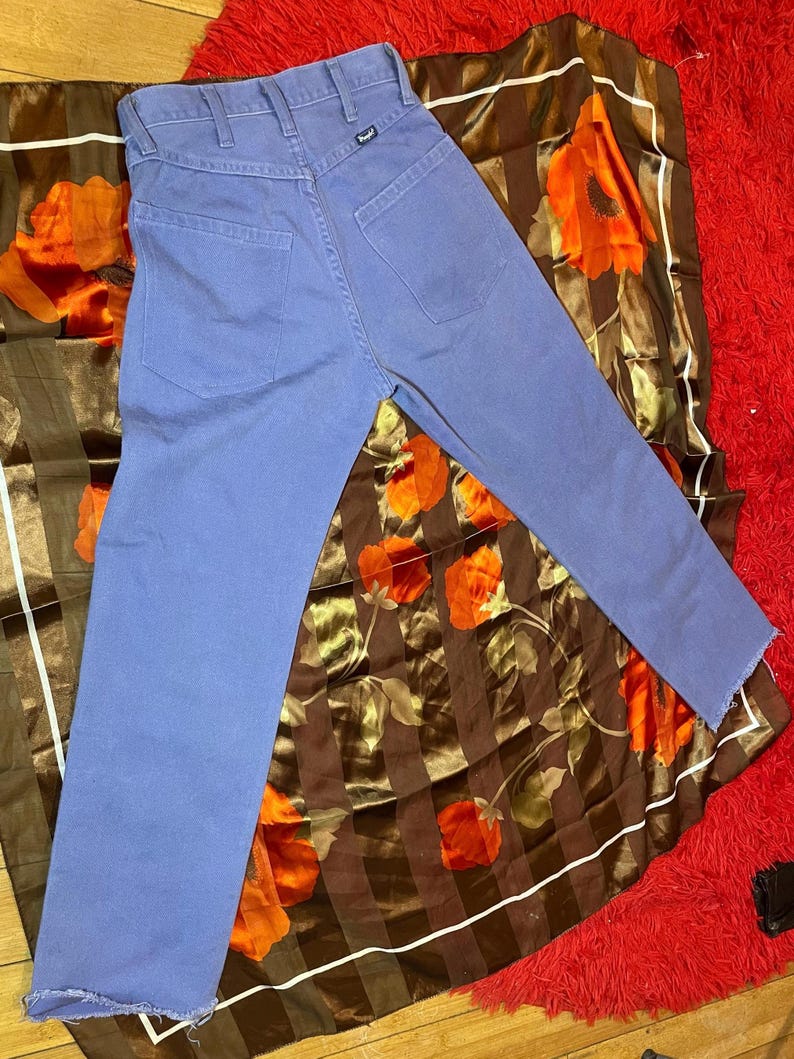 May include: A pair of blue jeans with a straight leg and frayed hem. The jeans are laid on a brown and orange floral patterned scarf, which is on top of a red rug.