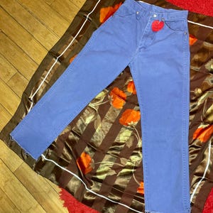 May include: Blue denim jeans with a red heart-shaped tag. The jeans are laid on a brown and orange floral patterned scarf, with a red rug visible in the background. The jeans have a classic five-pocket design.