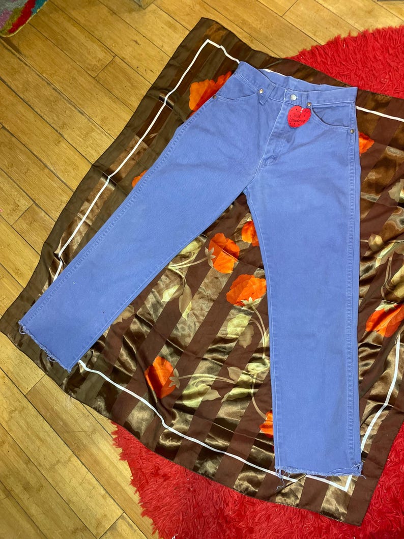 May include: Blue denim jeans with a red heart-shaped tag. The jeans are laid on a brown and orange floral patterned scarf, with a red rug visible in the background. The jeans have a classic five-pocket design.