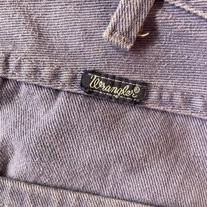 May include: Close-up of a section of light purple denim fabric. The Wrangler brand label is visible, stitched onto the denim. The fabric has a textured appearance with visible vertical lines.