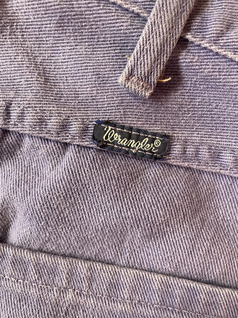 May include: Close-up of a section of light purple denim fabric. The Wrangler brand label is visible, stitched onto the denim. The fabric has a textured appearance with visible vertical lines.
