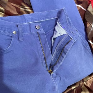 May include: Close-up of a pair of blue jeans with a partially unzipped zipper. The jeans have a button closure and pockets. The fabric appears to be a solid, medium-weight denim. The jeans are folded.