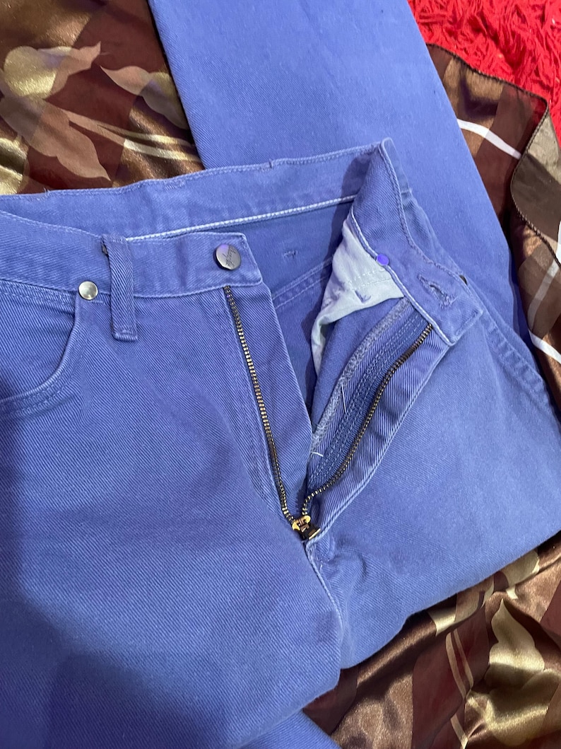 May include: Close-up of a pair of blue jeans with a partially unzipped zipper. The jeans have a button closure and pockets. The fabric appears to be a solid, medium-weight denim. The jeans are folded.