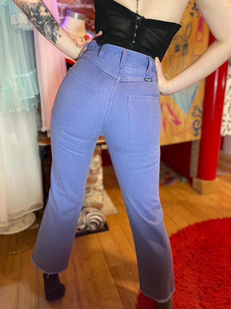 May include: Light blue denim jeans with a high waist and a slight flare at the hem. The jeans have a Wrangler label and classic five-pocket styling. The denim has a faded, ombre effect, transitioning to a darker shade at the bottom.
