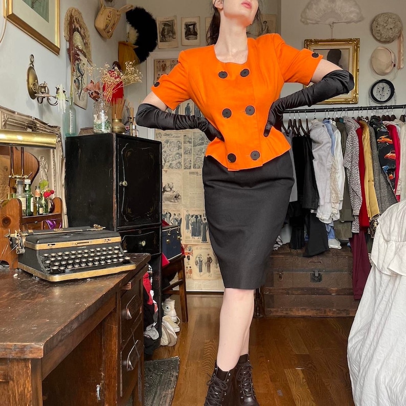May include: A woman wearing a bright orange short-sleeved jacket with black buttons and a black skirt. She is wearing black gloves and black boots.