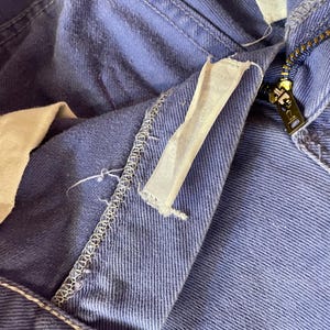 May include: Close-up of blue denim pants with a visible zipper and a white fabric tag. The pants show signs of wear, with visible stitching and a slightly faded color. The zipper is gold-toned.