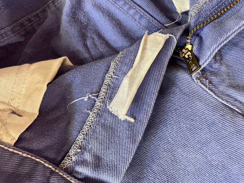 May include: Close-up of blue denim pants with a visible zipper and a white fabric tag. The pants show signs of wear, with visible stitching and a slightly faded color. The zipper is gold-toned.