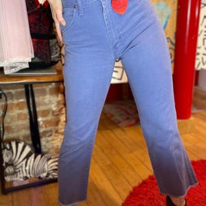 May include: Light blue denim jeans with a high waist and a slight flare at the ankle. The jeans have a faded appearance and a red heart-shaped tag attached to the front. The jeans are worn with black ankle boots.