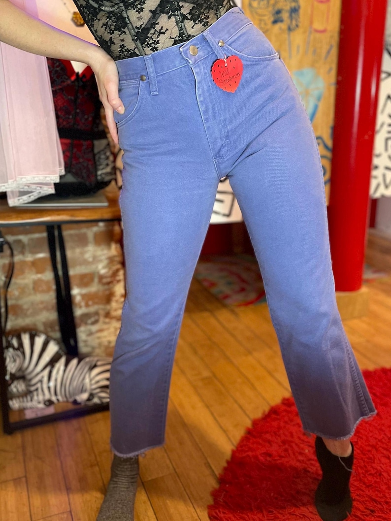May include: Light blue denim jeans with a high waist and a slight flare at the ankle. The jeans have a faded appearance and a red heart-shaped tag attached to the front. The jeans are worn with black ankle boots.