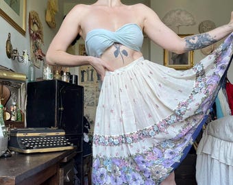 May include: A light blue tube top and a cream-colored skirt with a floral pattern. The skirt features a border of purple and blue flowers. The outfit is vintage-inspired and suitable for warm weather.