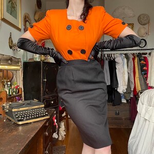 May include: A woman wearing a short-sleeved orange dress with black buttons and a black skirt. She is wearing black gloves and has her hair pulled back.