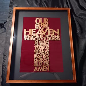 May include: Framed artwork featuring a gold-colored cross with the text of the Lord's Prayer. The cross is set against a burgundy background within a black frame, encased in a wooden frame.