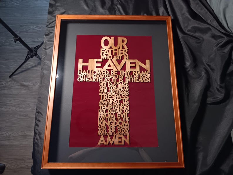 May include: Framed artwork featuring a gold-colored cross with the text of the Lord's Prayer. The cross is set against a burgundy background within a black frame, encased in a wooden frame.