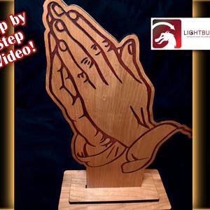May include: A wooden sculpture of praying hands, with detailed lines and a natural wood grain finish. The hands are depicted in a clasped position, mounted on a rectangular base. The image includes the text "Step by Step Video!" and a logo for Lightburn.