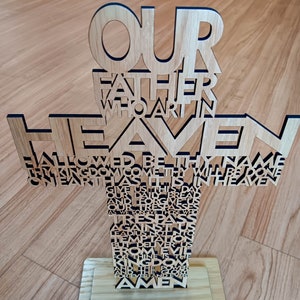 May include: Wooden cross with the text of the Lord's Prayer cut out. The cross is made of light-colored wood and stands on a rectangular base. The words "Our Father Who Art in Heaven" are at the top.