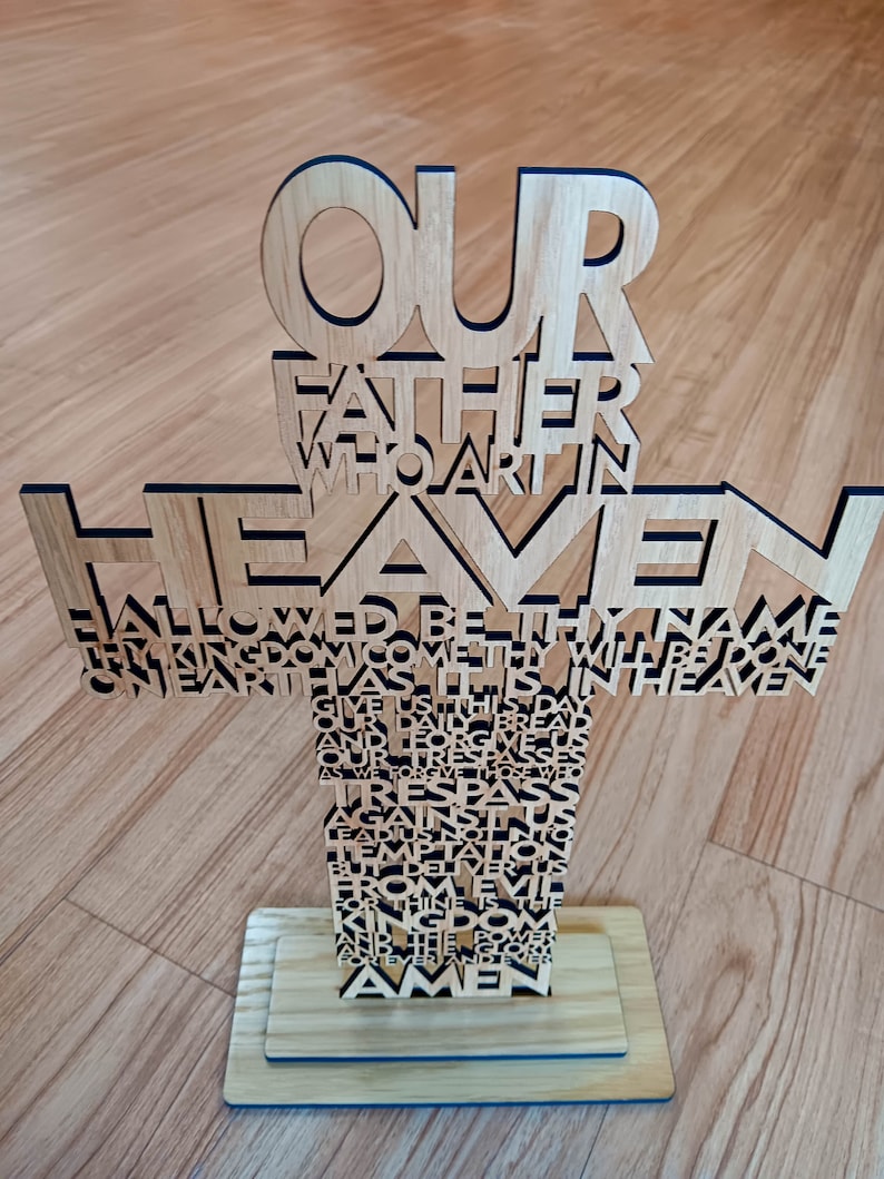 May include: Wooden cross with the text of the Lord's Prayer cut out. The cross is made of light-colored wood and stands on a rectangular base. The words "Our Father Who Art in Heaven" are at the top.