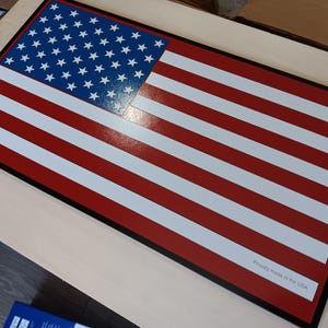 May include: A red, white, and blue American flag with a black border. The flag is made of wood and has a glossy finish. The text "Proudly made in the USA" is printed in the lower right corner.