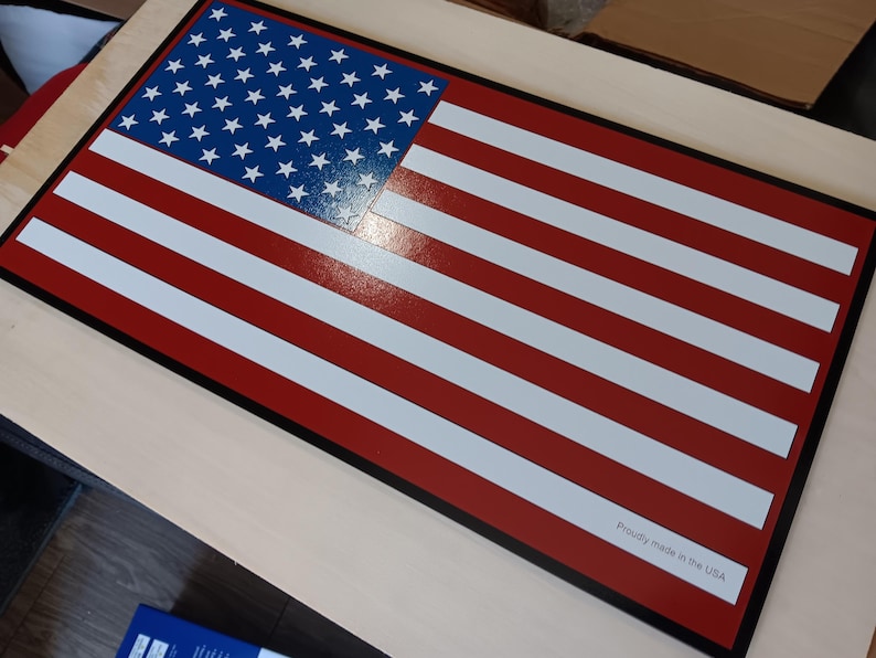 May include: A red, white, and blue American flag with a black border. The flag is made of wood and has a glossy finish. The text "Proudly made in the USA" is printed in the lower right corner.